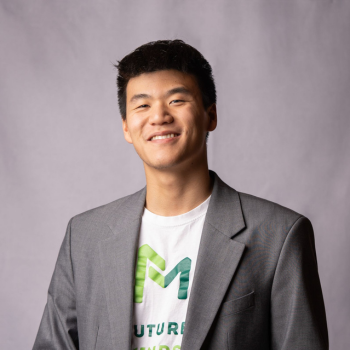 Smiling young man wearing a grey blazer and T-shirt against a plain background.
